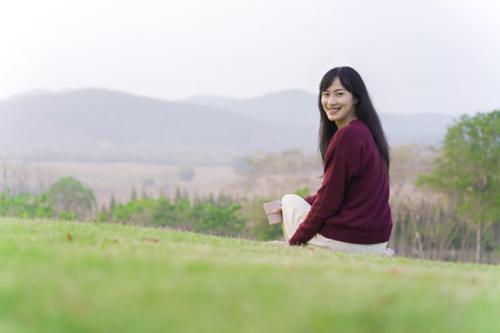 Happy beautiful Asian young woman sitting on the grass field and enjoy relaxing with a stunning view of mountain and forest landscape. Asian young woman drinking a cup of coffee in morning.の写真素材