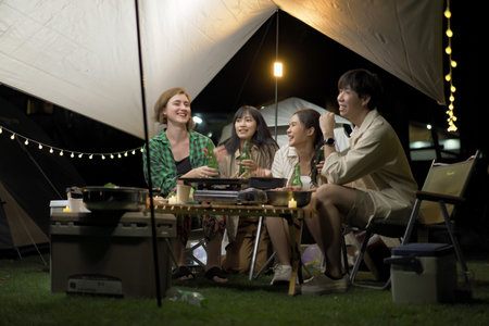 Group of diverse ethnicity young people enjoy eating and grilling bar-b-q together while camping in forest at night. Happy friends enjoy drinking a beer and playing   a guitar between camping.の写真素材