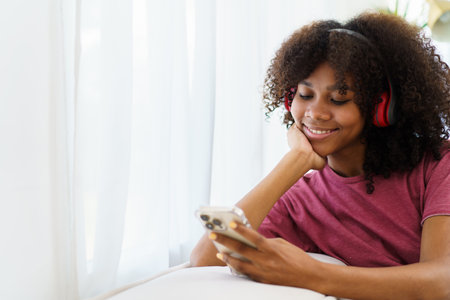 Happy cheerful American - African black teenager woman relaxing in the living room and using smartphone to browse the social network or internet. Woman relaxing in living room and using smartphone.の写真素材