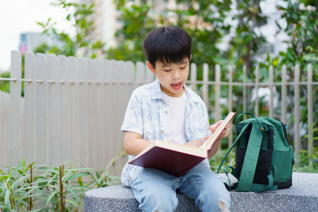 Cheerful happy Asian little boy sitting on the bench and reading a book or fairy tale at the public park alone.の写真素材