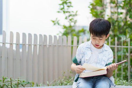 Cheerful happy Asian little boy sitting on the bench and reading a book or fairy tale at the public park alone.の写真素材