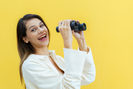 Beautiful caucasian white young woman looking through the twin lens binocular. Woman using binocular portrait on yellow background.の写真素材