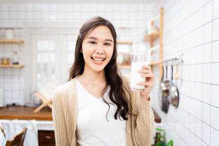 Beautiful happy Asian woman drinking a milk in morning in kitchen and smiling to camera. Happy Asian woman shows a glass of milk and smiles.の写真素材
