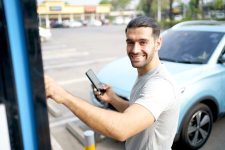 Cheerful happy Asian man using an EV charging application on smartphone to prepare vehicle charging and payment. Modern lifestyle of transportation with sustainability and sustainable energy.の写真素材