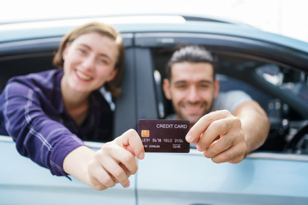 Happy cheerful young couple traveling by the car, male car driver showing a credit card to camera. Concept of travel and transportation insurance. Man paying a gas or EV charging by credit card.の写真素材
