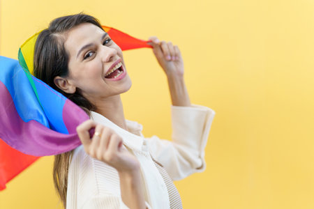 Happy cheerful caucasian white woman smiling and holding a rainbow flag which is the symbolic of LGBTQ people on yellow background. The diversity in genders and lifestyles concept.の写真素材