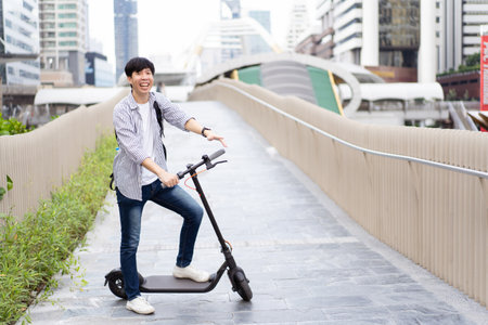 Happy cheerful Asian young man riding an electric scooter for traveling in the city area and street. New modern urban lifestyles concept. Happy young man smiles to camera while riding a scooter.の写真素材