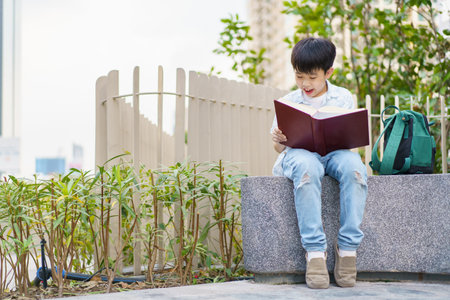 Cheerful happy Asian little boy sitting on the bench and reading a book or fairy tale at the public park alone.の写真素材