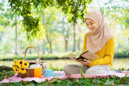 Happy cheerful Asian beautiful muslim woman sitting on the ground at the park and drinking a cup of tea in afternoon. Beautiful muslim woman relaxing and picnic in weekend.の写真素材