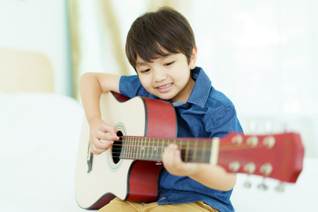 Happy cheerful Asian little young boy practices playing an acoustic guitar in bedroom. Lovely Asian little boy playing a guitar.の写真素材