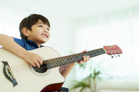 Happy cheerful Asian little young boy practices playing an acoustic guitar in bedroom. Lovely Asian little boy playing a guitar.の写真素材