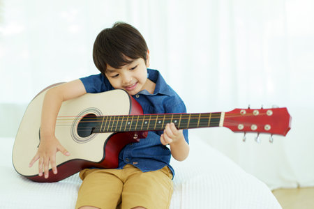 Happy cheerful Asian little young boy practices playing an acoustic guitar in bedroom. Lovely Asian little boy playing a guitar.の写真素材