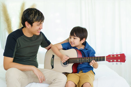 Asian little boy playing an acoustic guitar with his father, a man teaching and playing a guitar with his son in weekend. Happy family - fatherhood concept. Dad teaches his son play a guitar.の写真素材