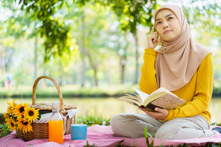Happy cheerful Asian beautiful muslim woman sitting on the ground at the park and drinking a cup of tea in afternoon. Beautiful muslim woman relaxing and picnic in weekend.の写真素材