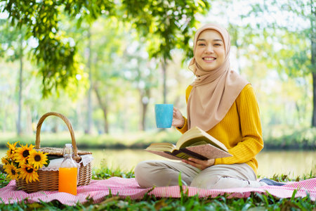 Happy cheerful Asian beautiful muslim woman sitting on the ground at the park and drinking a cup of tea in afternoon. Beautiful muslim woman relaxing and picnic in weekend.の写真素材