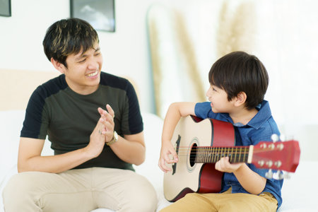 Asian little boy playing an acoustic guitar with his father, a man teaching and playing a guitar with his son in weekend. Happy family - fatherhood concept. Dad teaches his son play a guitar.の写真素材