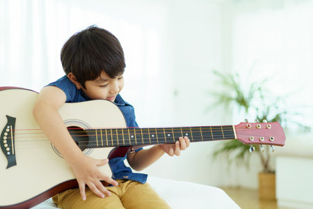 Happy cheerful Asian little young boy practices playing an acoustic guitar in bedroom. Lovely Asian little boy playing a guitar.の写真素材