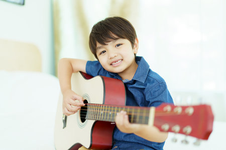 Happy cheerful Asian little young boy practices playing an acoustic guitar in bedroom. Lovely Asian little boy playing a guitar.の写真素材