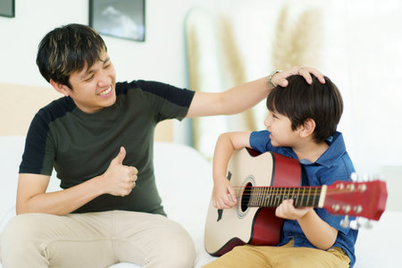 Asian little boy playing an acoustic guitar with his father, a man teaching and playing a guitar with his son in weekend. Happy family - fatherhood concept. Dad teaches his son play a guitar.の写真素材