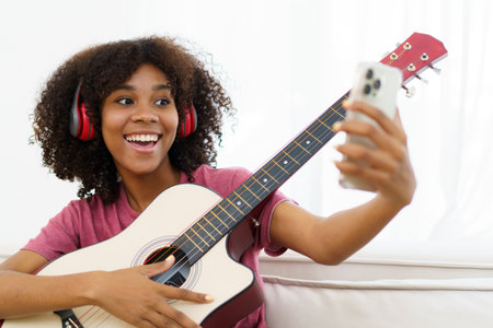 Happy cheerful African - American black woman playing in living room and making a live video or steaming on social media by using smartphone. Woman performs a guitar and makes live video steaming.の写真素材