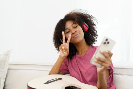 Happy cheerful African - American black woman playing in living room and making a live video or steaming on social media by using smartphone. Woman performs a guitar and makes live video steaming.の写真素材