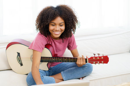 Happy cheerful American - African black woman sitting on a sofa in living room and playing an acoustic guitar close up with copy space.の写真素材