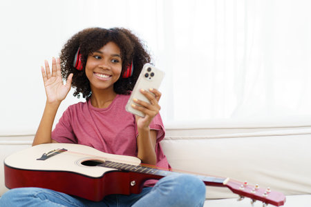 Happy cheerful African - American black woman playing in living room and making a live video or steaming on social media by using smartphone. Woman performs a guitar and makes live video steaming.の写真素材