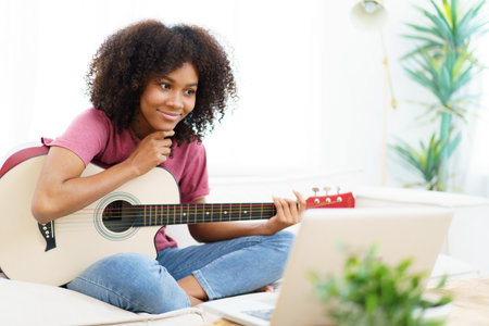 Happy cheerful American - African black woman sitting on a sofa in living room and playing an acoustic guitar close up with copy space.の写真素材