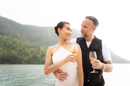 Happy Asian and caucasian white couple celebrating their marriage anniversary together on the yacht. Adult couple sailing a yacht and drinking a Champaign together during a honeymoon trip.の写真素材
