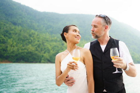 Happy Asian and caucasian white couple celebrating their marriage anniversary together on the yacht. Adult couple sailing a yacht and drinking a Champaign together during a honeymoon trip.の写真素材