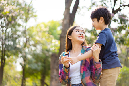 Asian woman and boy playing and walking around the park together with beautiful bokeh and lighting background copyspace. Little cute boy hogging his mother from back and kissing.の写真素材