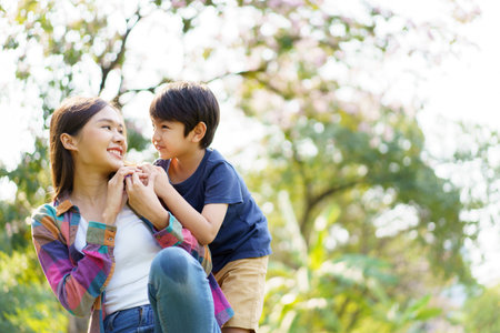 Asian woman and boy playing and walking around the park together with beautiful bokeh and lighting background copyspace. Little cute boy hogging his mother from back and kissing.の写真素材