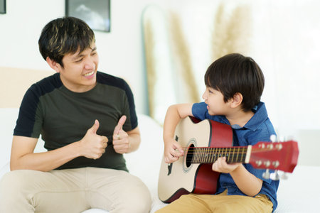 Asian little boy playing an acoustic guitar with his father, a man teaching and playing a guitar with his son in weekend. Happy family - fatherhood concept. Dad teaches his son play a guitar.の写真素材
