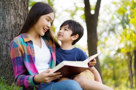 Happy cheerful Asian lovely little boy and mother sitting under the tree at park in the morning and reading a book together. Asian woman spending time in weekend with her lovely little son.の写真素材