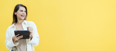 Happy cheerful caucasian white female student holding a digital tablet and taking a note on tablet on yellow background. Smart and beautiful woman student posing on yellow background.の写真素材