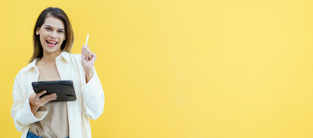Happy cheerful caucasian white female student holding a digital tablet and taking a note on tablet on yellow background. Smart and beautiful woman student posing on yellow background.の写真素材