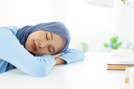 Asian young female muslim student feels sleepy and yawn while doing her homework at home. Lazy - bored muslim woman sitting in the home office and yawning.の写真素材