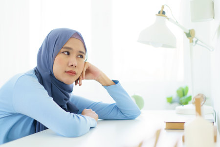 Asian young female muslim student feels sleepy and yawn while doing her homework at home. Lazy - bored muslim woman sitting in the home office and yawning.の写真素材