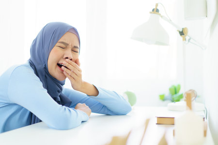 Asian young female muslim student feels sleepy and yawn while doing her homework at home. Lazy - bored muslim woman sitting in the home office and yawning.の写真素材