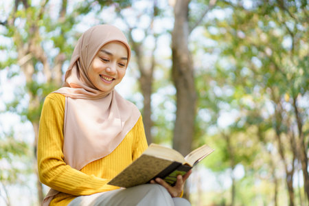 Happy cheerful Asian beautiful Muslim woman reading a book - Koran under the tree. Female muslim student studying an Islam Koran.の写真素材