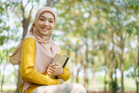 Happy cheerful Asian beautiful Muslim woman reading a book - Koran under the tree. Female muslim student studying an Islam Koran.の写真素材