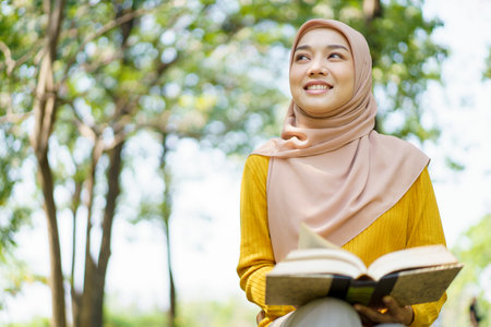Happy cheerful Asian beautiful Muslim woman reading a book - Koran under the tree. Female muslim student studying an Islam Koran.の写真素材