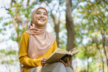 Happy cheerful Asian beautiful Muslim woman reading a book - Koran under the tree. Female muslim student studying an Islam Koran.の写真素材