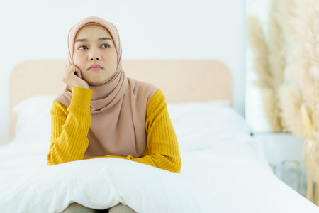 Unhappy - stressful Asian young muslim woman sitting alone on the bed in the morning. Asian muslim woman having a mental burn out or depression problem from working and social environment.の写真素材