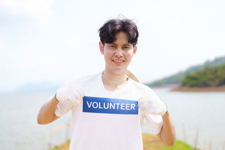 Handsome Asian volunteer smiling to camera portrait, man pointing finger at his volunteer t-shirt. Happy cheerful male volunteer proudly smiles to camera while working.の写真素材