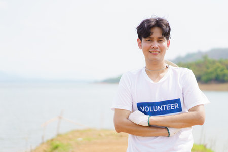 Handsome Asian volunteer smiling to camera portrait, man pointing finger at his volunteer t-shirt. Happy cheerful male volunteer proudly smiles to camera while working.の写真素材