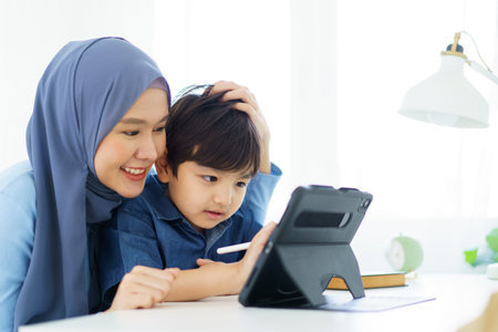 Happy cheerful Asian muslim woman woman teaching her little son a drawing on digital tablet, mother let her little young boy doing a school homework or an online learning on tablet.の写真素材
