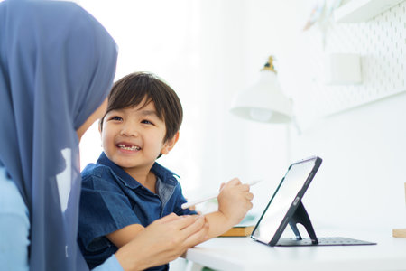 Happy cheerful Asian muslim woman woman teaching her little son a drawing on digital tablet, mother let her little young boy doing a school homework or an online learning on tablet.の写真素材