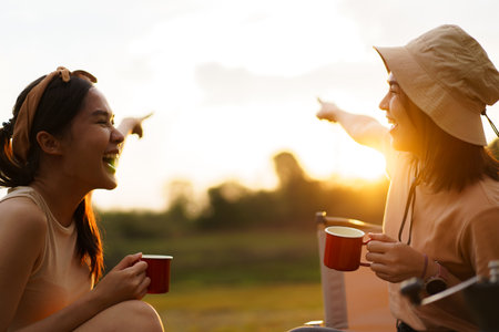 Happy cheerful Asian young women - friends camping in the lake forest and enjoy drinking a cup of tea or coffee. Female friends on a camping trip in the natural park in summer.の写真素材