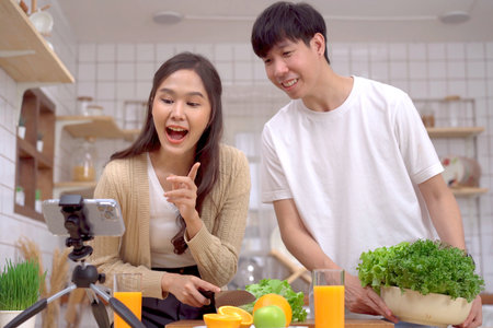 Happy cheerful Asian man and woman talking about cooking and food nutrition in a beautiful decorated kitchen on social network together. Lovely couple making an online live steaming on social media.の写真素材